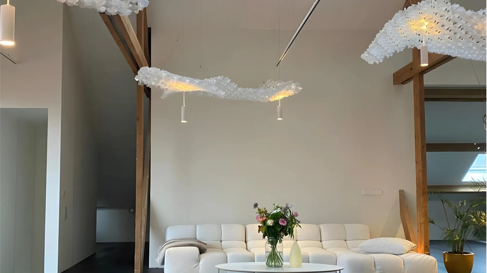 Woonkamer met zwevende lichtobjecten onder houten spanten, een witte bank en bloemen op tafel